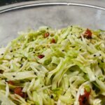 A vibrant cabbage salad in a large wooden bowl with chopped green cabbage, sliced red tomatoes, green onions, and black sesame seeds, garnished with parsley leaves and a drizzle of sesame oil. Text overlay reads: 'How to Make a Delicious Cabbage Salad at Home | Easy & Healthy Recipe.