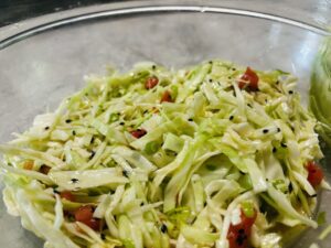 A vibrant cabbage salad in a large wooden bowl with chopped green cabbage, sliced red tomatoes, green onions, and black sesame seeds, garnished with parsley leaves and a drizzle of sesame oil. Text overlay reads: 'How to Make a Delicious Cabbage Salad at Home | Easy & Healthy Recipe.